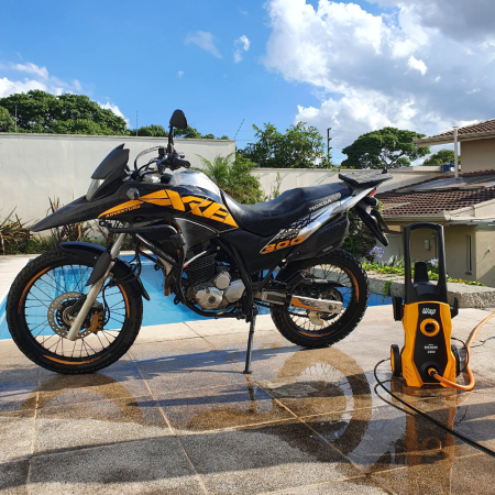 Motocicleta preta com detalhes em amarelo apoiada no descanso lateral sobre piso molhado de cerâmica, ao lado direito uma lavadora de alta pressão amarela
