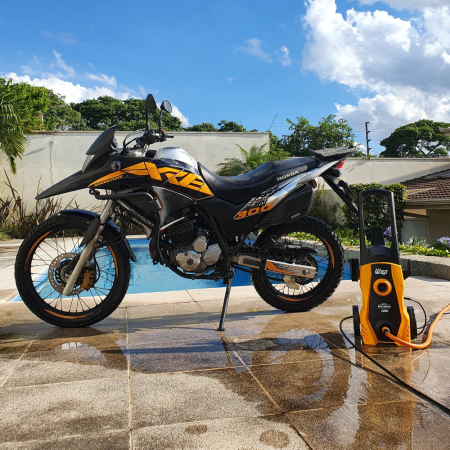 Motocicleta preta com detalhes em amarelo estacionada sobre piso molhado ao lado de uma lavadora amarela, em um pátio com piscina ao fundo