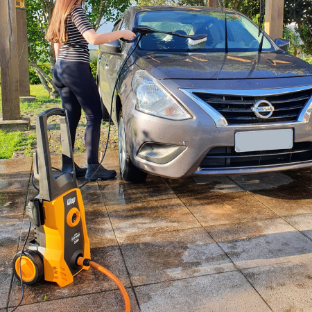 Mulher de perfil, vestindo calça preta e blusa listrada, usando uma lavadora amarela para limpar a parte frontal de um carro Nissan cinza