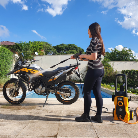 Mulher sorridente, vestindo blusa listrada e calça preta, usando a pistola de alta pressão de uma lavadora amarela para lavar a parte traseira de uma motocicleta