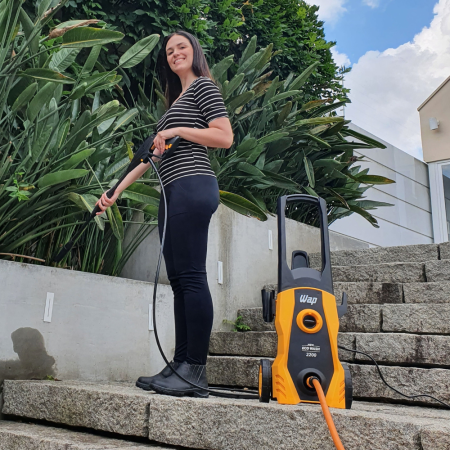 Mulher sorridente, vestindo calça preta e blusa listrada, em pé sobre degraus de pedra, usando uma lavadora de alta pressão WAP amarela para limpar a superfície;
